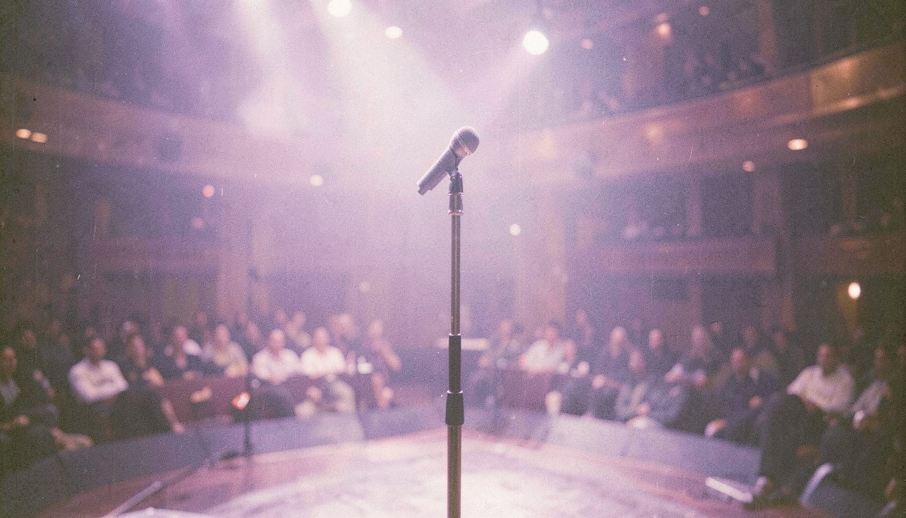 Yumuşak mor ve krem tonlarında, loş bir sahne ışığı altında boş bir mikrofon standı; arka planda flu bir konser salonu atmosferi.