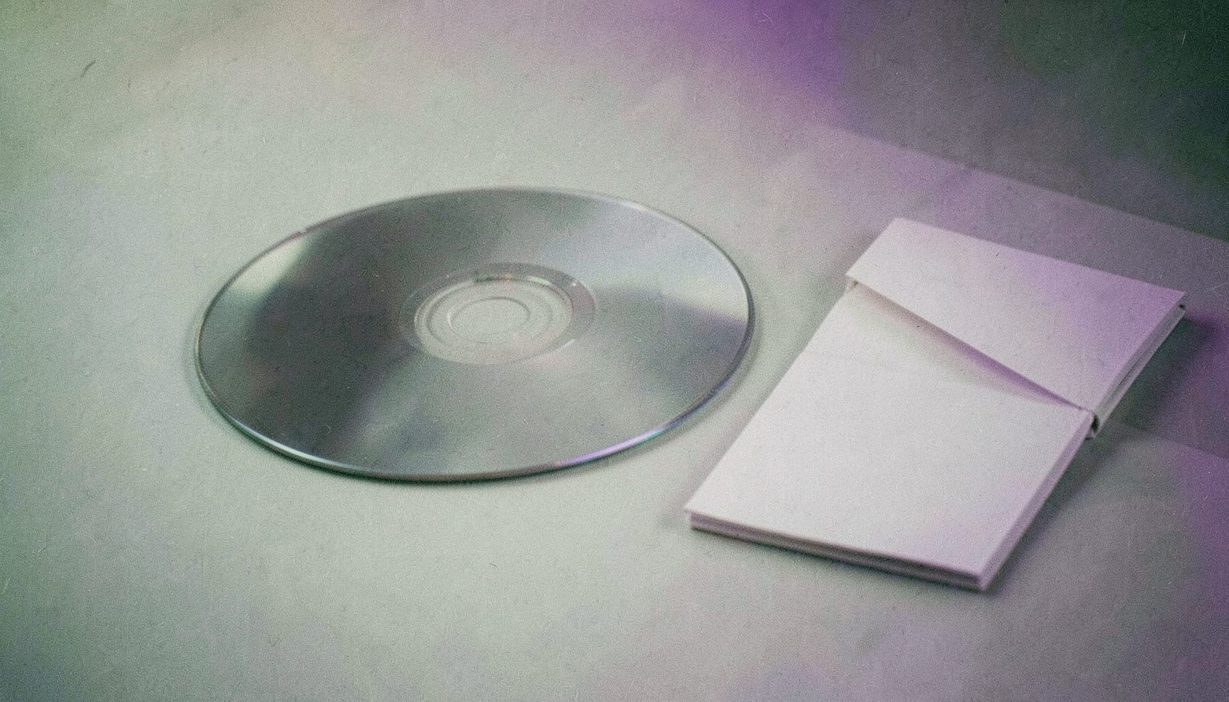 Sıcak krem ve yumuşak yeşil tonlarında, düz bir yüzey üzerinde duran bir CD ve yanında katlanmış bir kitapçık; yumuşak gölgeler ve film greniyle sakin bir kompozisyon.