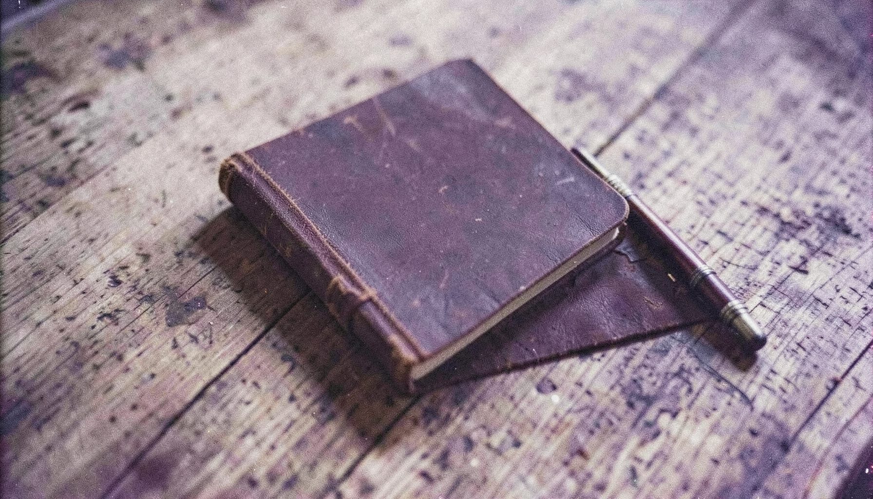 Koyu kahve ve krem tonlarında, rustik bir ahşap zemin üzerinde duran deri kaplı bir defter ve yanında eski bir kalem; analog film dokusuyla zamansız bir atmosfer.