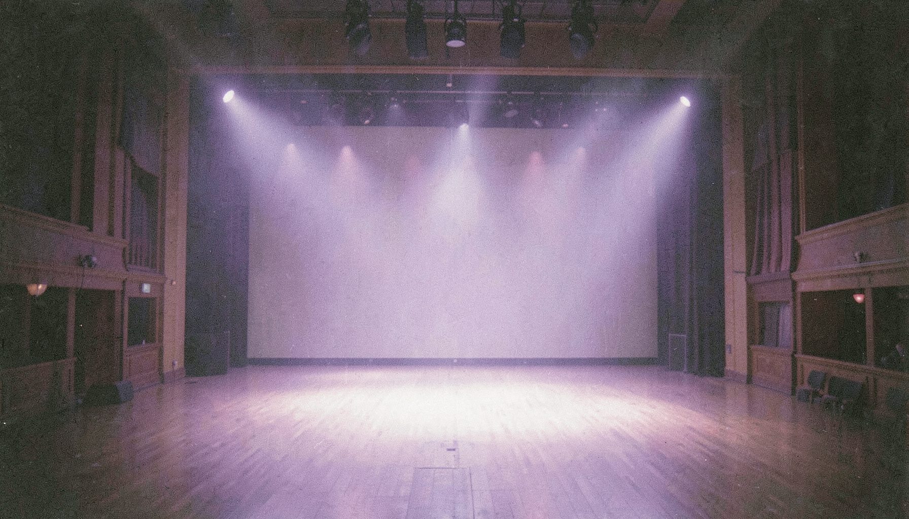 Yumuşak gri ve lavanta tonlarında, bir konser salonunun boş sahnesini gösteren geniş açılı bir kare; sahne ışıkları loş, analog film greniyle melankolik ve sanatsal bir hava.